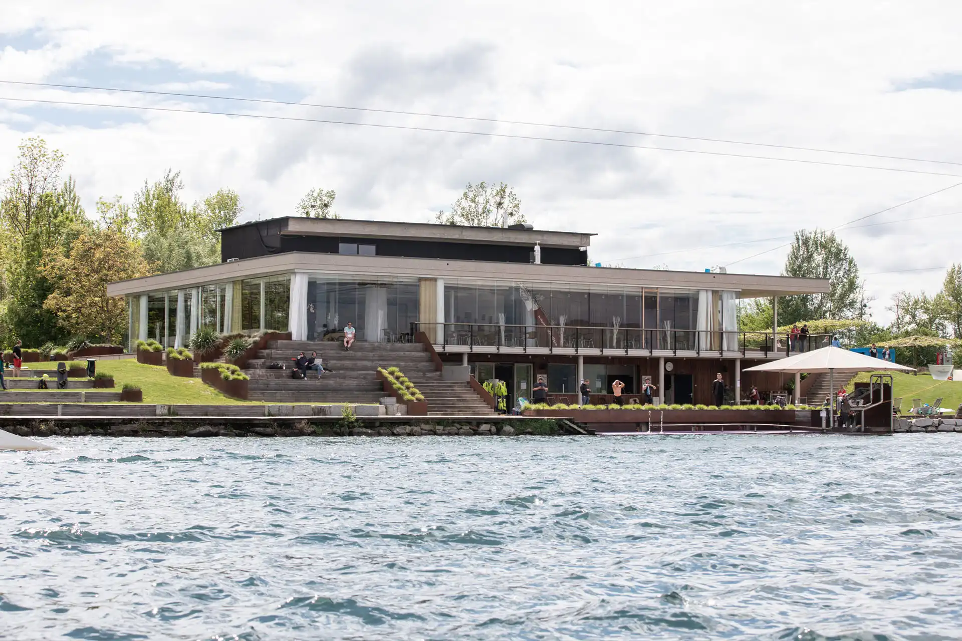 Ein modernes Gebäude mit Glaswänden und Flachdach steht am Wasser, umgeben von grünem Gras und Bäumen. Mehrere Personen entspannen sich auf den Stufen und im Terrassenbereich, und im Vordergrund ist das Wasser zu sehen.
