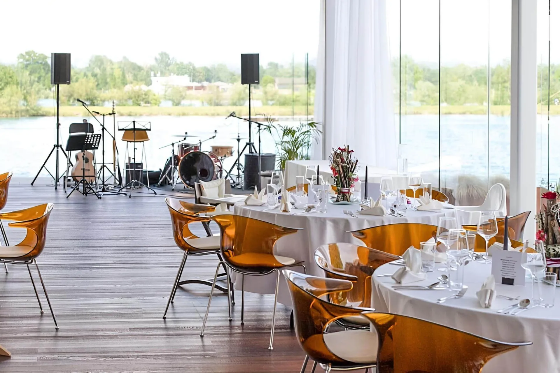 Ein moderner Veranstaltungsraum mit großen Fenstern, runden Tischen zum Essen, bernsteinfarbenen, durchsichtigen Stühlen und einer Bühne mit Musikinstrumenten und Mikrofonen, von der aus man einen schönen Blick ins Freie hat.