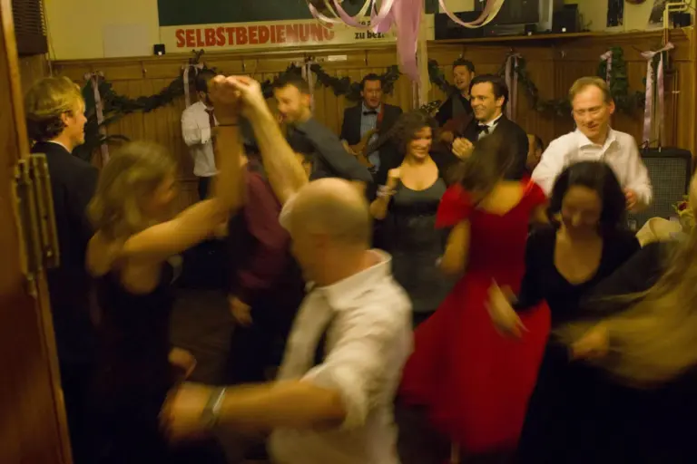 Eine Gruppe von formell gekleideten Personen tanzt energisch in einem warm beleuchteten Raum, während im Hintergrund Musiker spielen. Von der Decke hängen rosa Luftschlangen, und an der Wand ist ein SELBSTBEDIENUNG-Schild zu sehen.