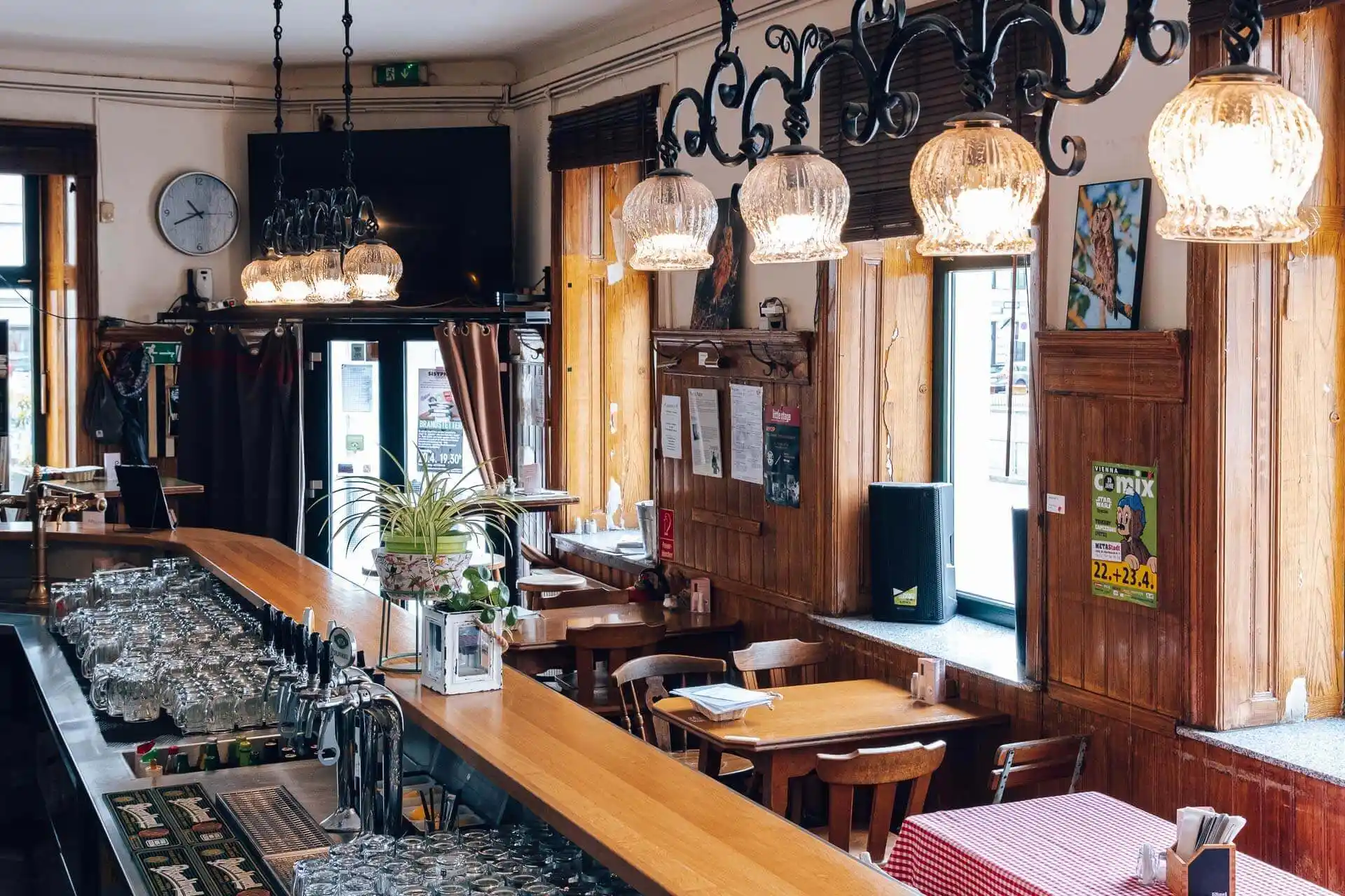 Eine gemütliche, gut beleuchtete Café-Bar mit Holztischen und -stühlen, einer langen Theke mit Gläsern, Hängelampen, großen Fenstern und Postern an den holzgetäfelten Wänden.