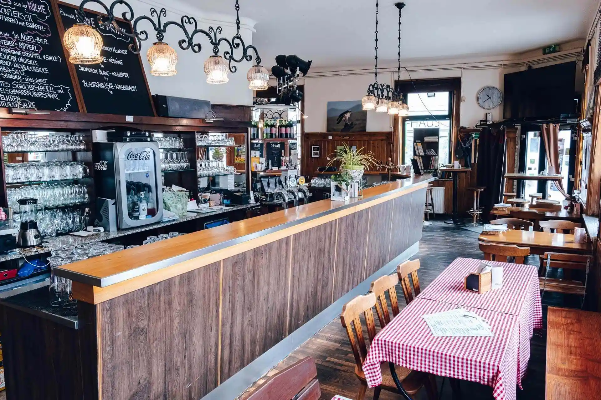 Ein gemütliches Café mit einer langen Holzbar, Hängelampen und Regalen mit Gläsern und Flaschen. Es gibt Holztische und -stühle, einen Tisch mit einer rot karierten Tischdecke und Speisekarten sowie große Fenster, die Tageslicht hereinlassen.
