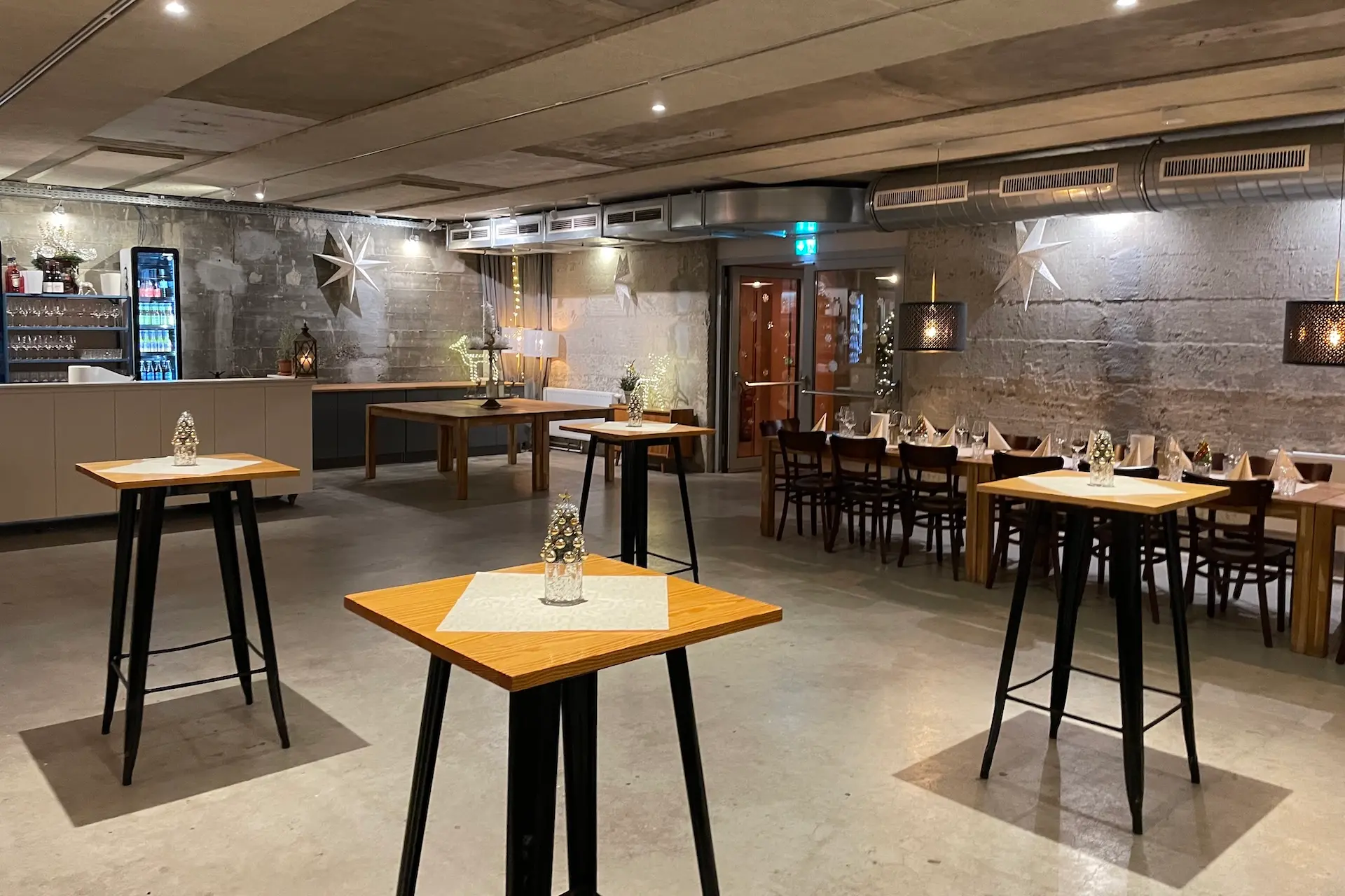 Ein moderner Veranstaltungsraum im Industrial-Stil mit Stehtischen, einem langen, für die Gäste gedeckten Esstisch und sanfter Beleuchtung. Dekorative Sterne und Glasflaschen sorgen für eine festliche Atmosphäre.