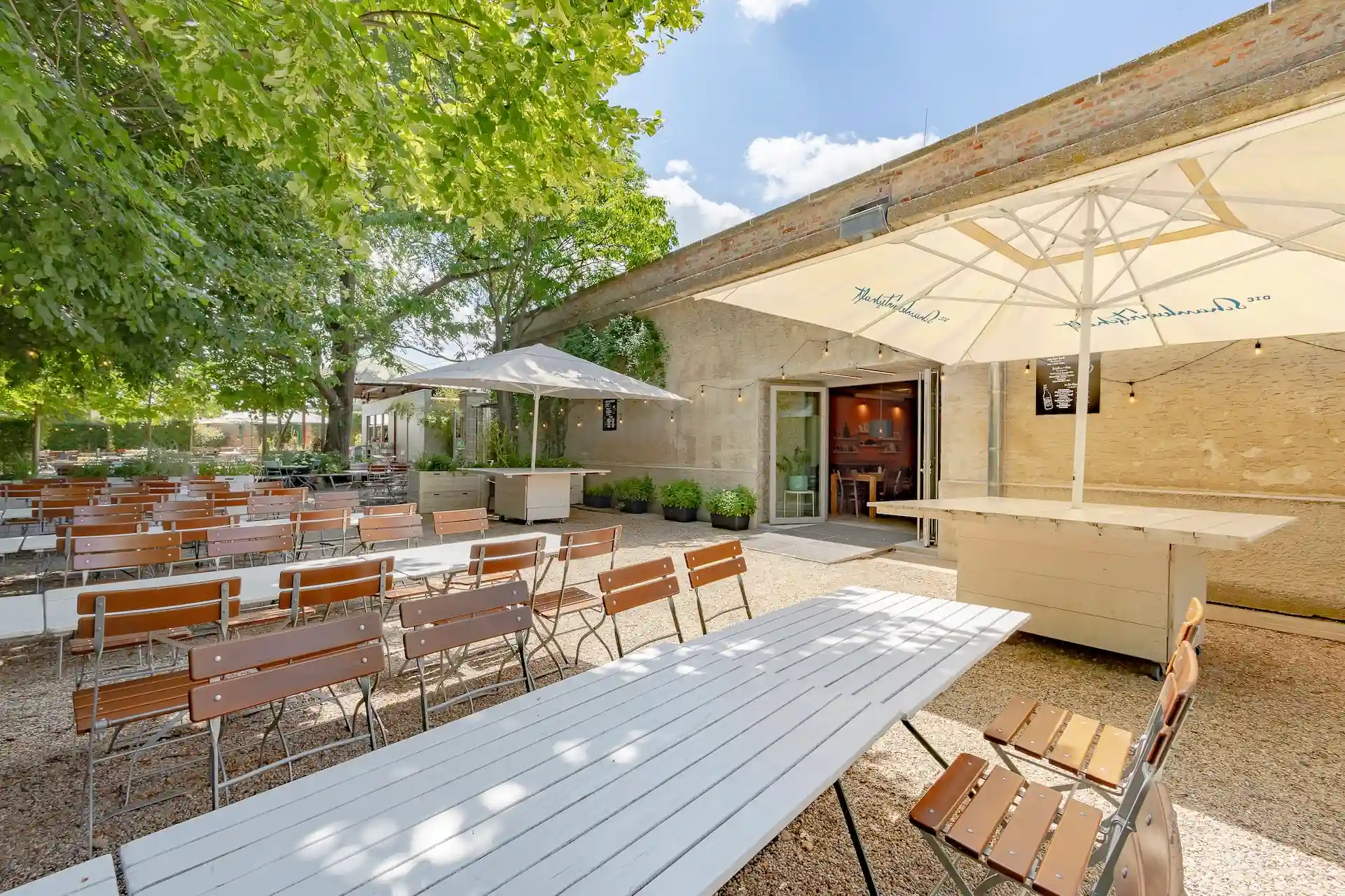 Sitzbereich im Freien mit Holztischen und -stühlen auf Kies, im Schatten großer Sonnenschirme und Bäume, neben einem beigen Gebäude mit offenen Türen unter strahlend blauem Himmel.
