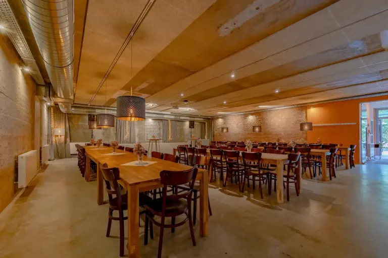 Geräumiger Speisesaal mit Holztischen und -stühlen, unverputzten Ziegel- und Betonwänden, Deckenleuchten und großen Hängelampen. Für eine Gruppenveranstaltung oder ein Beisammensein hergerichtet.
