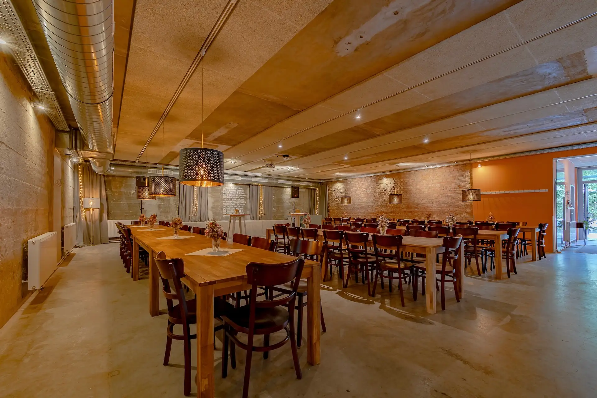 Geräumiger Speisesaal mit Holztischen und -stühlen, unverputzten Ziegel- und Betonwänden, Deckenleuchten und großen Hängelampen. Für eine Gruppenveranstaltung oder ein Beisammensein hergerichtet.