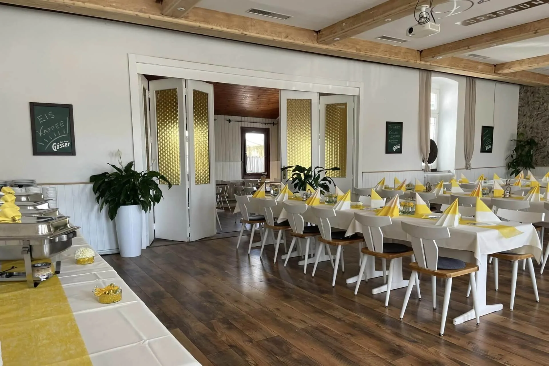 Ein Bankettsaal mit gelb und weiß gedeckten Tischen für eine Mahlzeit. Jeder Tisch hat gefaltete gelbe Servietten, weiße Stühle und kleine Pflanzen. Auf der einen Seite stehen Tische mit einem Buffet, an den Wänden befinden sich große Topfpflanzen und Kreidetafeln mit Speisekarten.
