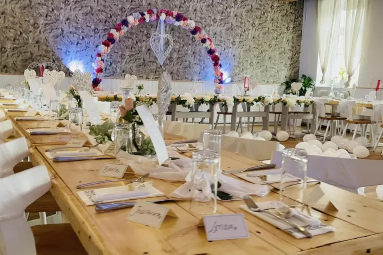 Ein langer, dekorierter Banketttisch ist für eine formelle Veranstaltung gedeckt, mit Namensschildern, Gläsern, Besteck und Blumensträußen. Im Hintergrund sind ein Blumenbogen und weitere Tische zu sehen, die auf eine Hochzeit oder Feier hindeuten.