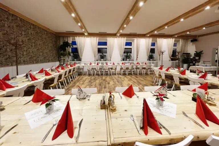 Ein geräumiger, gut beleuchteter Speisesaal, der für eine Veranstaltung eingerichtet ist, mit U-förmig angeordneten Holztischen. Auf jedem Platz liegen rote, zu Dreiecken gefaltete Servietten, weiße Stühle und kleine Blumenarrangements. Weiße Vorhänge verdecken die hohen Fenster.