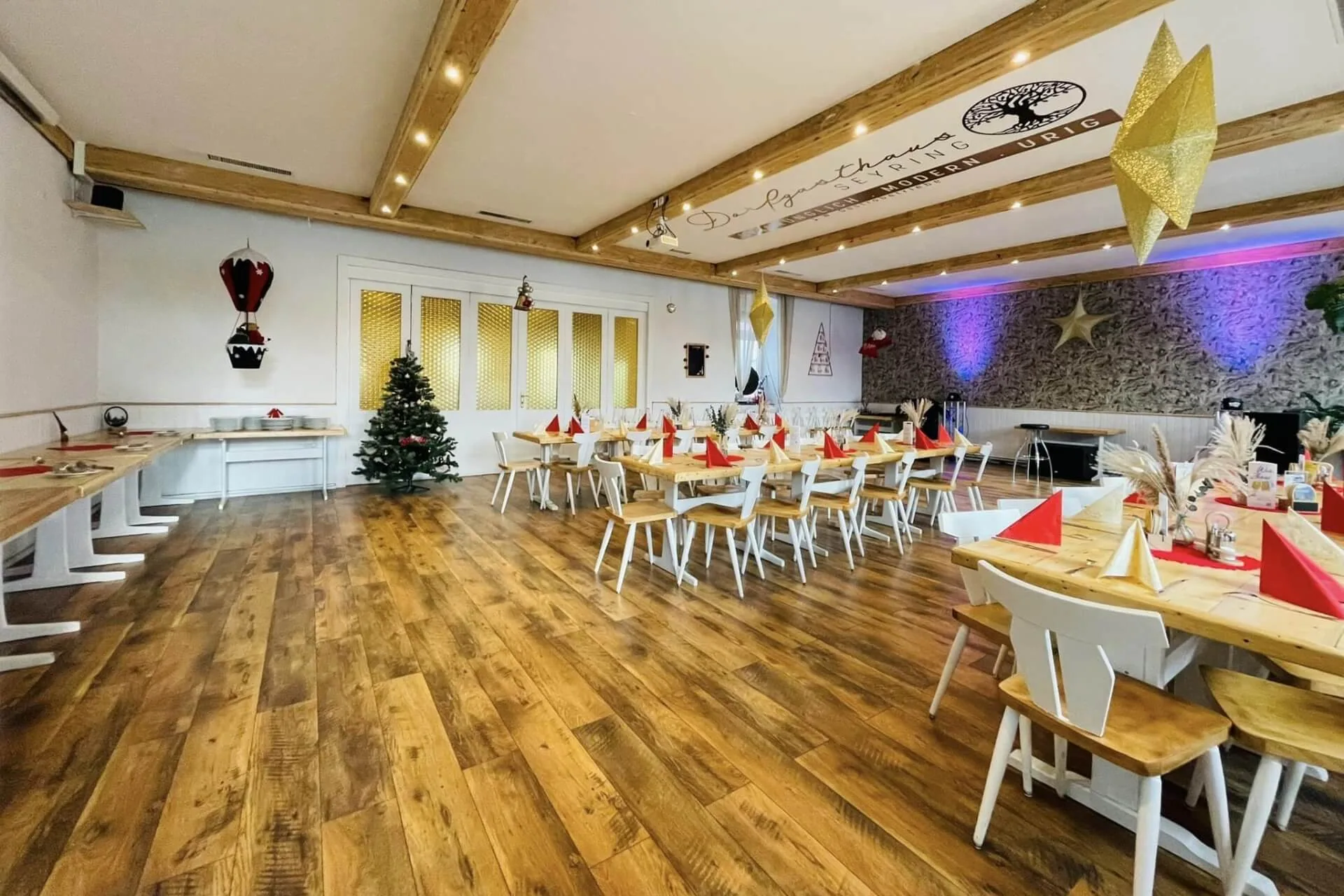 Ein festlicher Speisesaal mit Holzfußboden, weihnachtlich geschmückt mit einem Baum, goldenen Sternen und roten und gelben Servietten auf Tischen mit weißen Stühlen. Warme Beleuchtung und rustikale Balken schaffen eine gemütliche Atmosphäre.