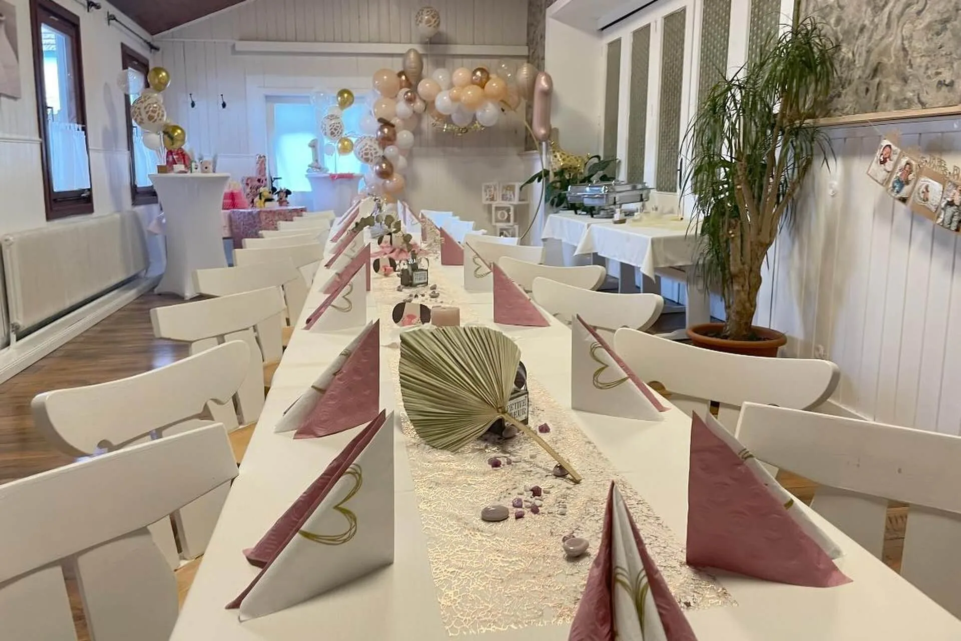 Ein langer Esstisch ist für eine Party gedeckt, mit gefalteten rosa und weißen Servietten, dekorativen Tafelaufsätzen und Luftballons im Hintergrund. Der Raum ist hell mit weißen Wänden und Holzböden.