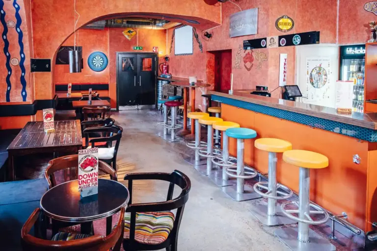Eine farbenfrohe Bar mit orangefarbenen Wänden, eine Reihe von Hockern mit bunten Sitzen am Tresen, Dartscheiben an der Wand, Holztische und -stühle sowie verschiedene Dekorationen und Schilder im ganzen Raum.