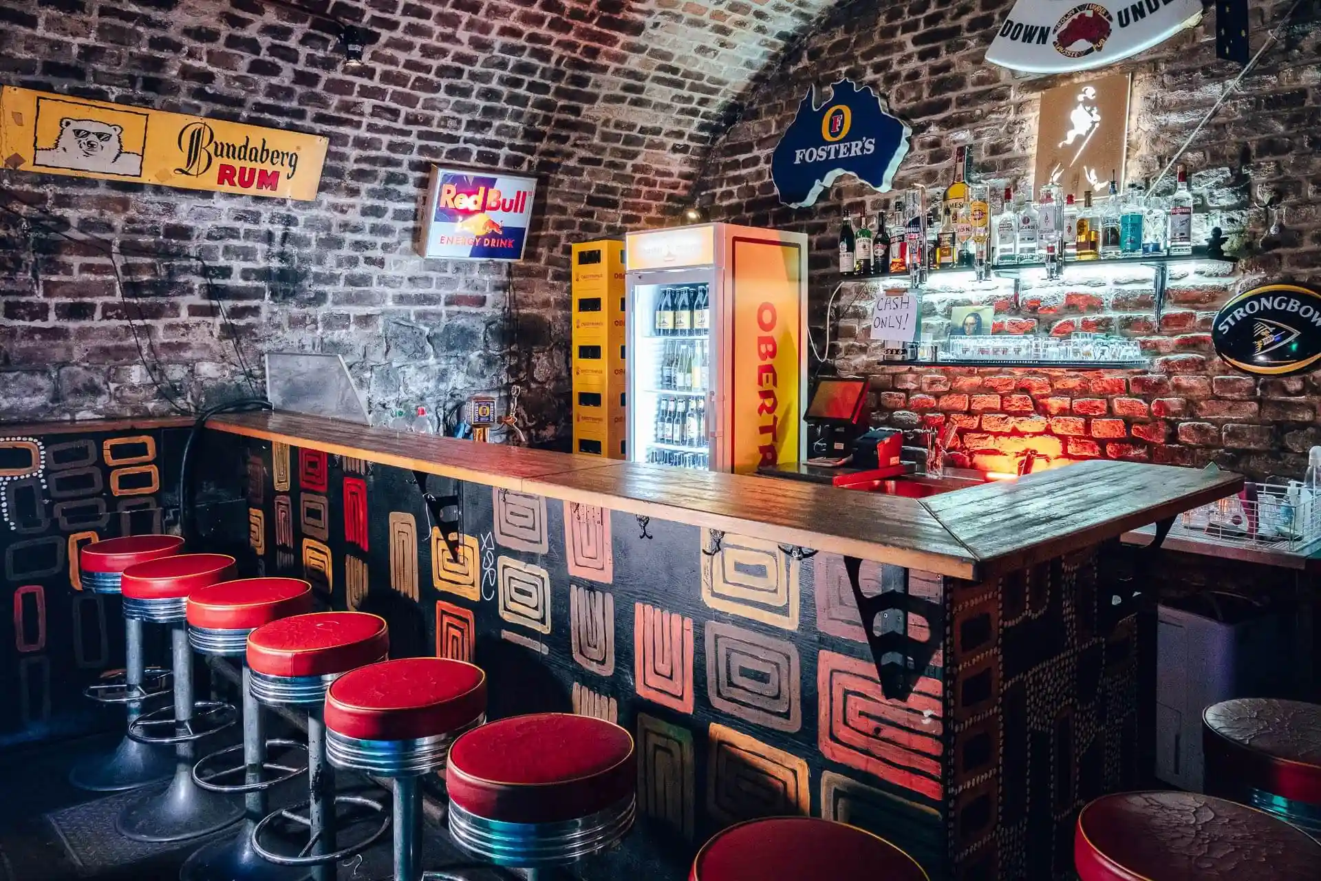 Eine gemütliche Bar mit rot gepolsterten Hockern, einem gemusterten Holztresen und freigelegten Backsteinwänden, die mit alten Schildern verziert sind. Hinter der Theke stehen Kühlschränke mit Getränken und Regale mit Flaschen und Gläsern.