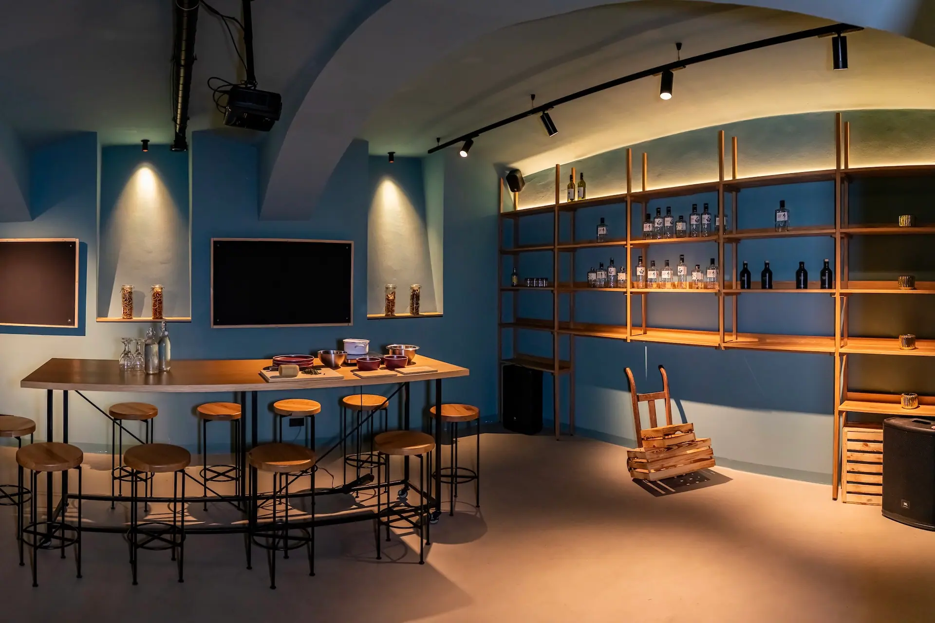 Drei Bögen Eine moderne Bar mit blauen Wänden, Holzregalen mit Flaschen, Barhockern um einen langen Holztresen und Scheinwerfern, die eine gemütliche Atmosphäre schaffen. In der Nähe der Regale ist ein Holzkarren umgekippt.