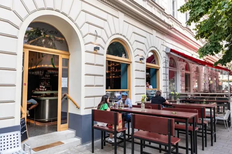 Menschen sitzen an Holztischen vor einem Café mit Bogenfenstern und hellen Wänden. Der Eingang ist offen und gibt den Blick auf das Innere frei, und Bäume spenden Schatten auf dem Gehweg.