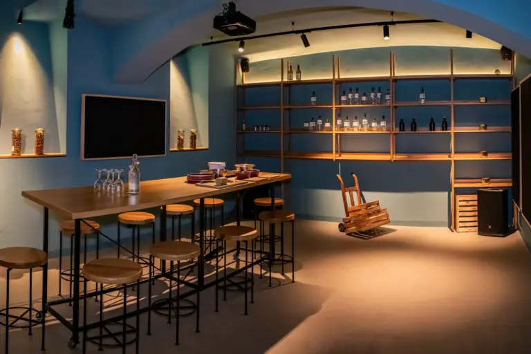 Ein moderner Barbereich mit Holzhockern um einen hohen Tisch, Regale mit Flaschen und Gläsern an einer blauen Wand und ein kleiner Holzwagen auf dem Boden bei warmer Beleuchtung.
