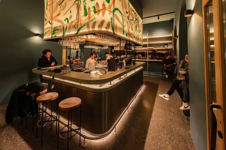 Moderne Bar mit geschwungenem Tresen, hohen Hockern und sitzenden oder stehenden Gästen. Warme Beleuchtung, hängende Weingläser und farbenfrohes abstraktes Deckendekor schaffen eine gemütliche, einladende Atmosphäre. Im hinteren Bereich sind Regale und Küche zu sehen.