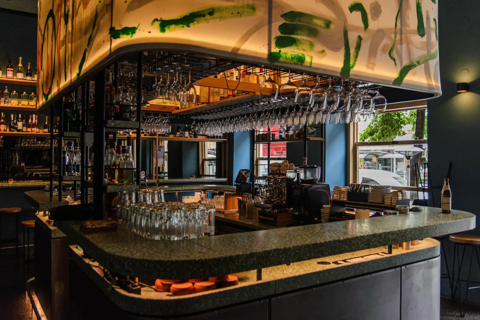 Drei Bögen Eine moderne Bar mit einer grün gesprenkelten Theke, hängenden Weingläsern, Regalen voller Flaschen und großen Fenstern, durch die Tageslicht hereinströmt.