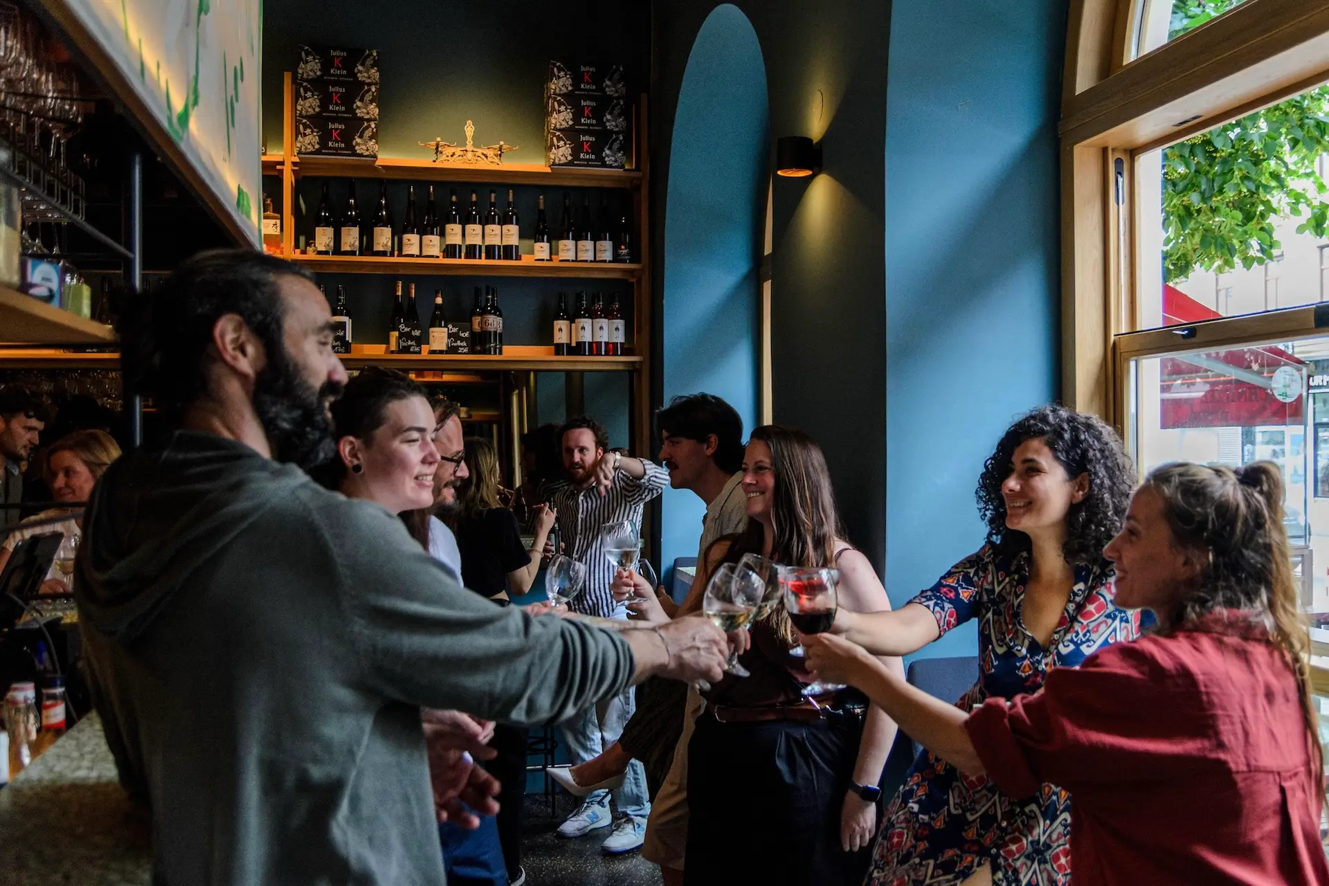 Eine Gruppe von Menschen steht in einer gemütlichen Bar, lächelt und stößt mit den Gläsern an – in der Nähe großer Fenster, durch die Tageslicht hereinströmt, umgeben von Regalen voller Flaschen und warmer Beleuchtung.