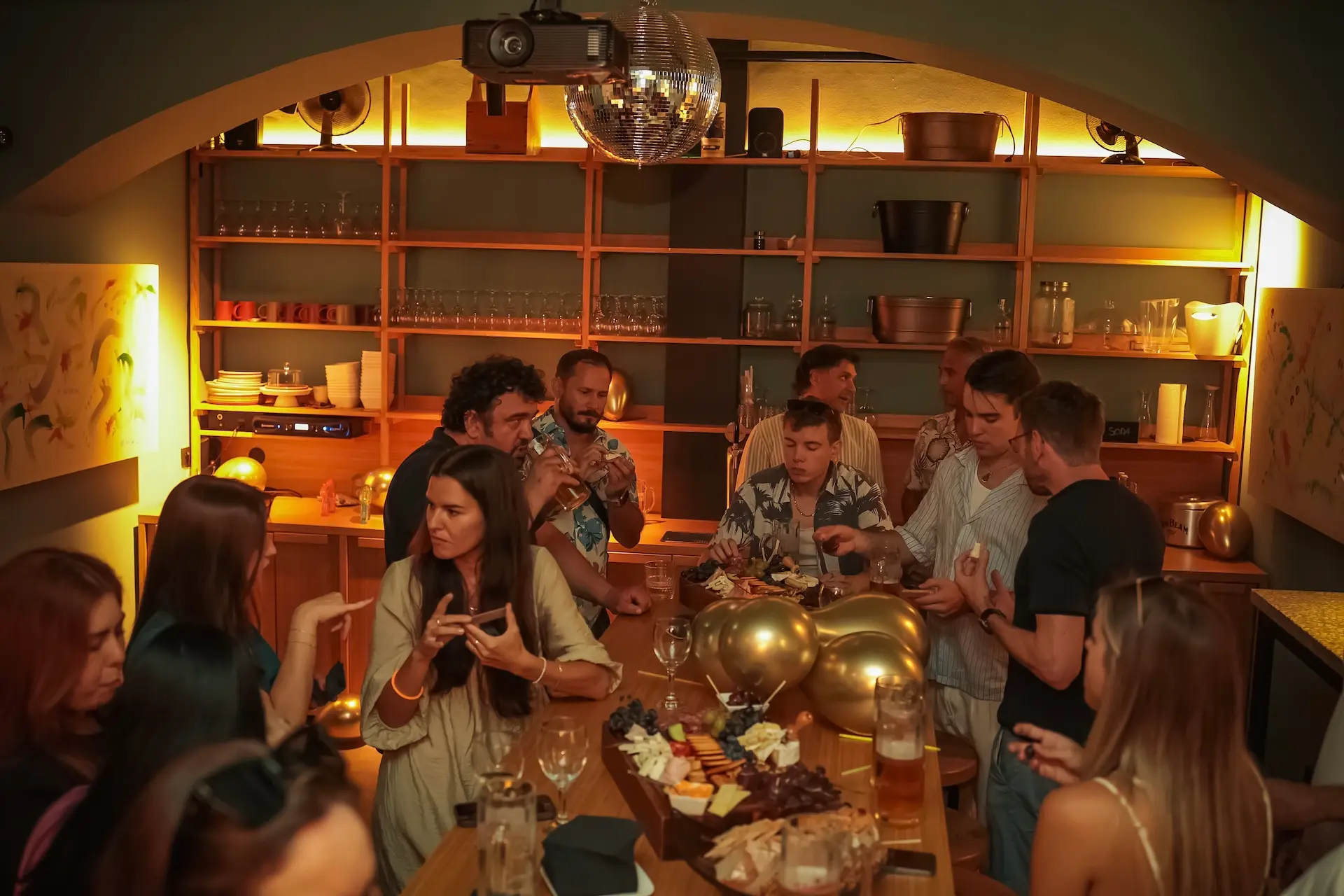 Eine Gruppe von Menschen versammelt sich in einer warm beleuchteten Bar mit Holzregalen und genießt Getränke und Speisen um einen mit Luftballons, Käse und Obsttellern dekorierten Tisch in der Mitte.