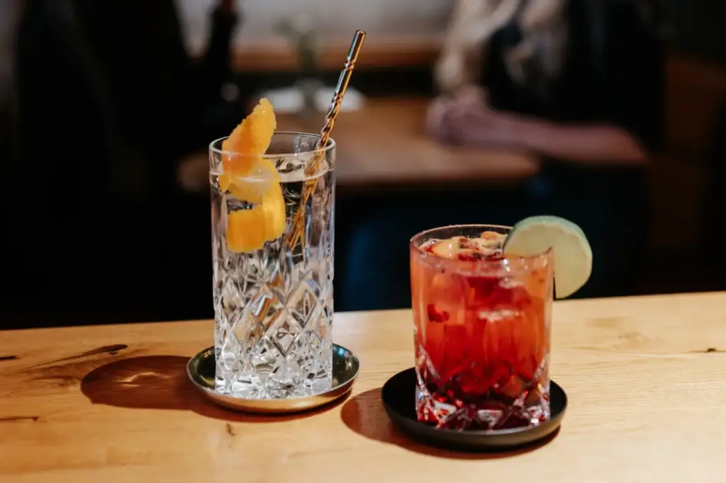 Zwei Kristallgläser auf einem Holztisch im Café Bar Laby: eines mit einem klaren Getränk, Eis, Orangenschale und einem goldenen Strohhalm; das andere mit einem roten Cocktail, Eis und einer Limettenscheibe. Im Hintergrund sitzen unscharfe Menschen.