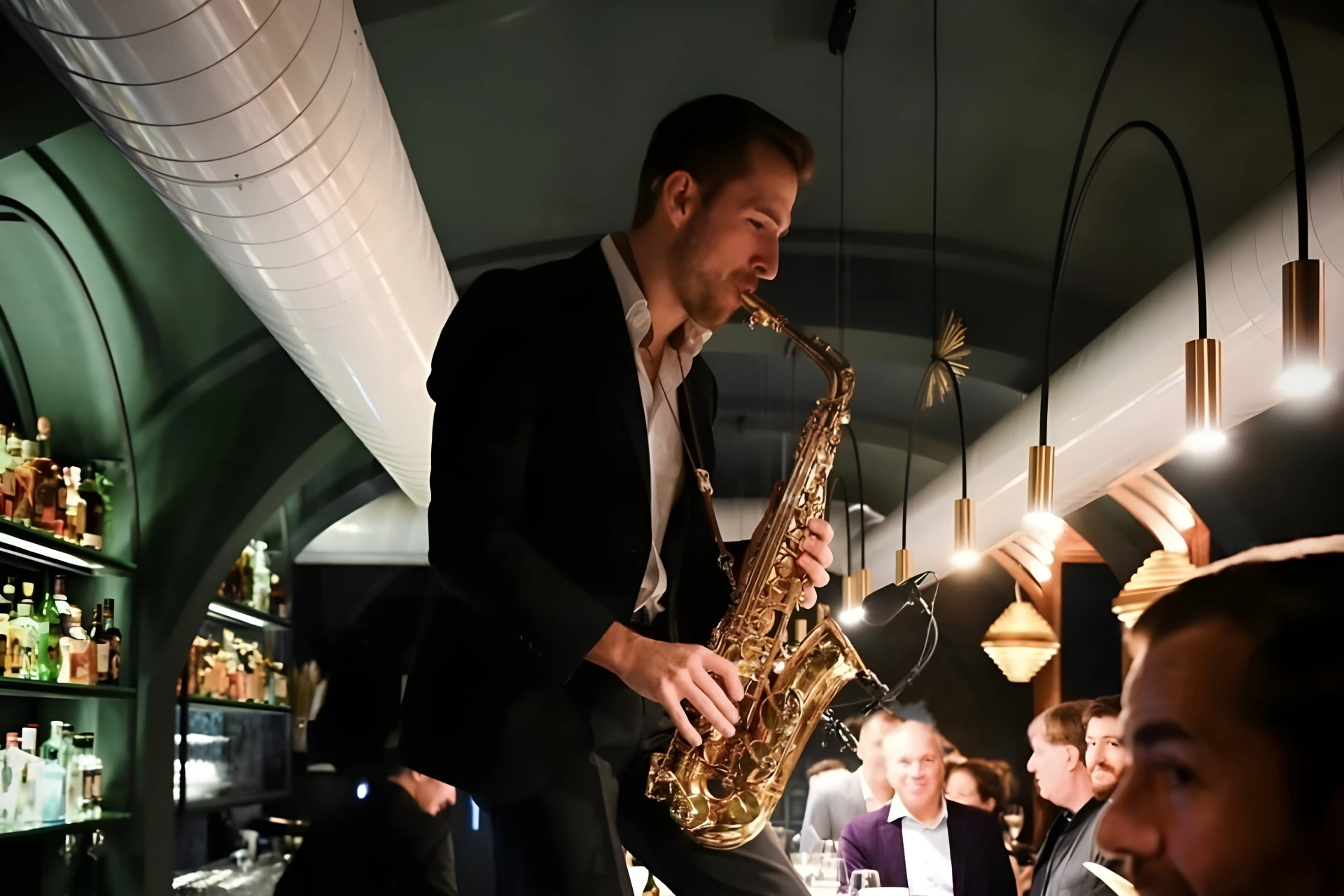 Ein Mann im Anzug spielt Saxophon, während er auf einem Bartresen in einem stilvollen, schwach beleuchteten Restaurant steht, in dem die Leute sitzen und ihn beobachten.