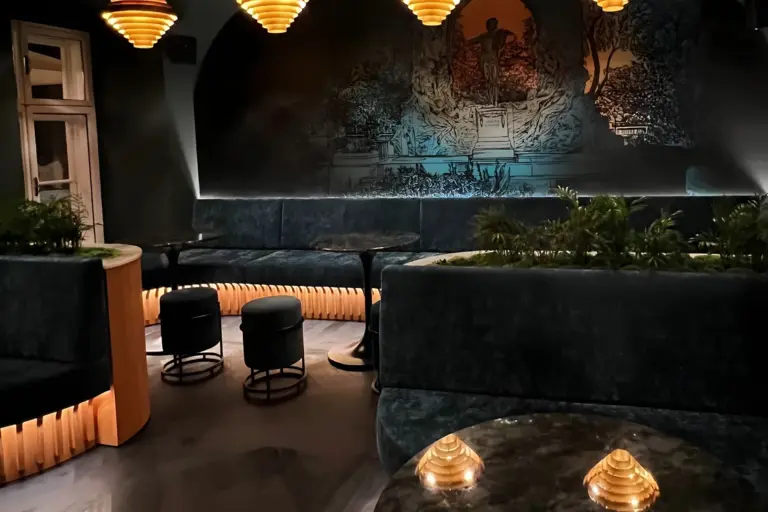 Eine schummrig beleuchtete Lounge mit dunklen Samtsesseln, runden Tischen, gepolsterten Hockern, goldenen Hängelampen und dekorativem Grün, mit einem Wandgemälde an der Rückwand und einer gemütlichen, modernen Atmosphäre.