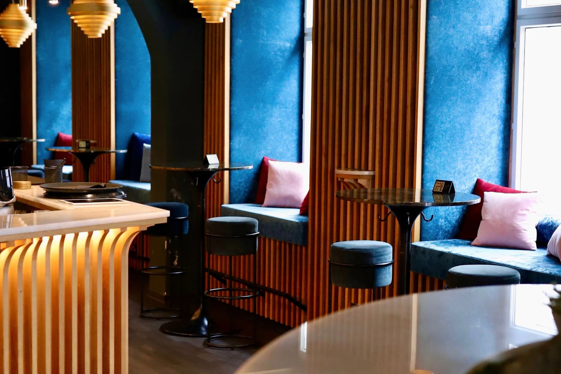 Ein modernes Café mit blauen Samtbestuhlung, Holzlattenwänden, runden Tischen, Barhockern und Hängelampen. Kissen in Rosa, Blau und Weiß akzentuieren die Fensterbänke.