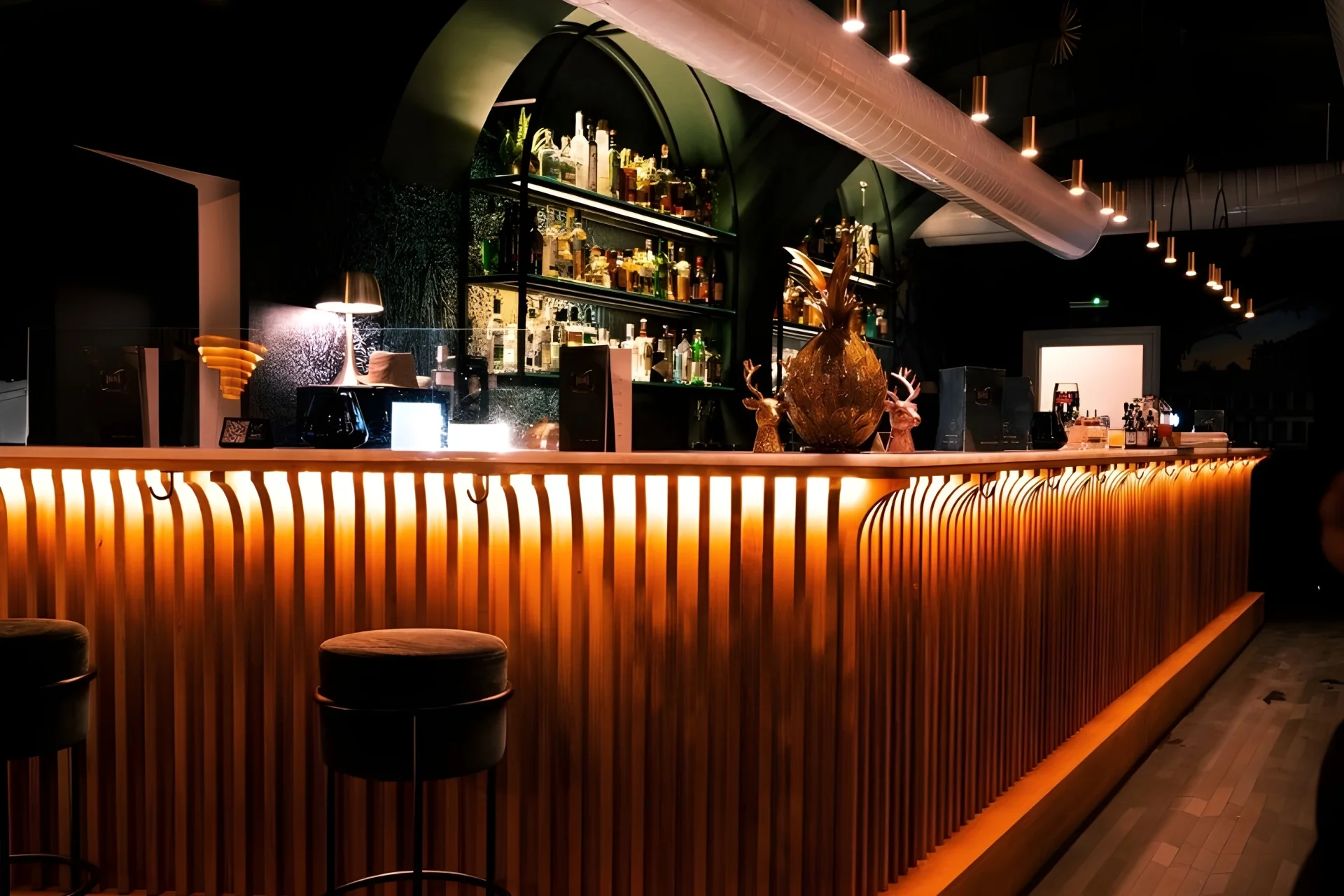 Eine moderne Bar mit einem hölzernen Tresen, der von warmen Lichtern beleuchtet wird, dahinter Regale mit Flaschen und Gläsern, davor ein einzelner Barhocker und eine stilvolle, einladende Atmosphäre.