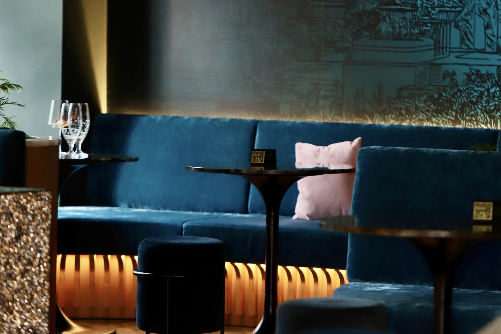 Eine stilvolle Lounge mit Sitzen aus blauem Samt, rosa Kissen, schwarzen Tischen mit Reservierungsschildern, leeren Weingläsern und sanfter, warmer Beleuchtung. Die Einrichtung ist modern und einladend.