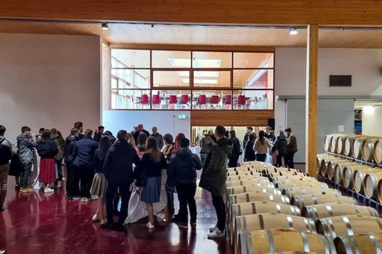Eine Gruppe von Menschen versammelt sich in einer Weinkellerei und steht auf einem roten Boden in der Nähe von Reihen von Weinfässern, wobei im Hintergrund der obere Bereich mit Glaswänden zu sehen ist.