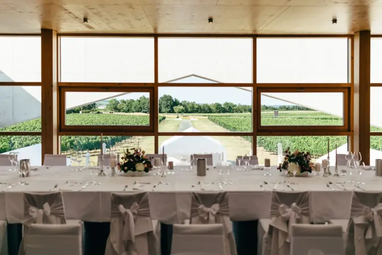 Ein langer, elegant gedeckter Tisch mit weißen Stühlen und Blumenschmuck steht im Innenbereich vor großen Fenstern, die einen Blick auf einen üppig grünen Weinberg unter einem klaren Himmel bieten.