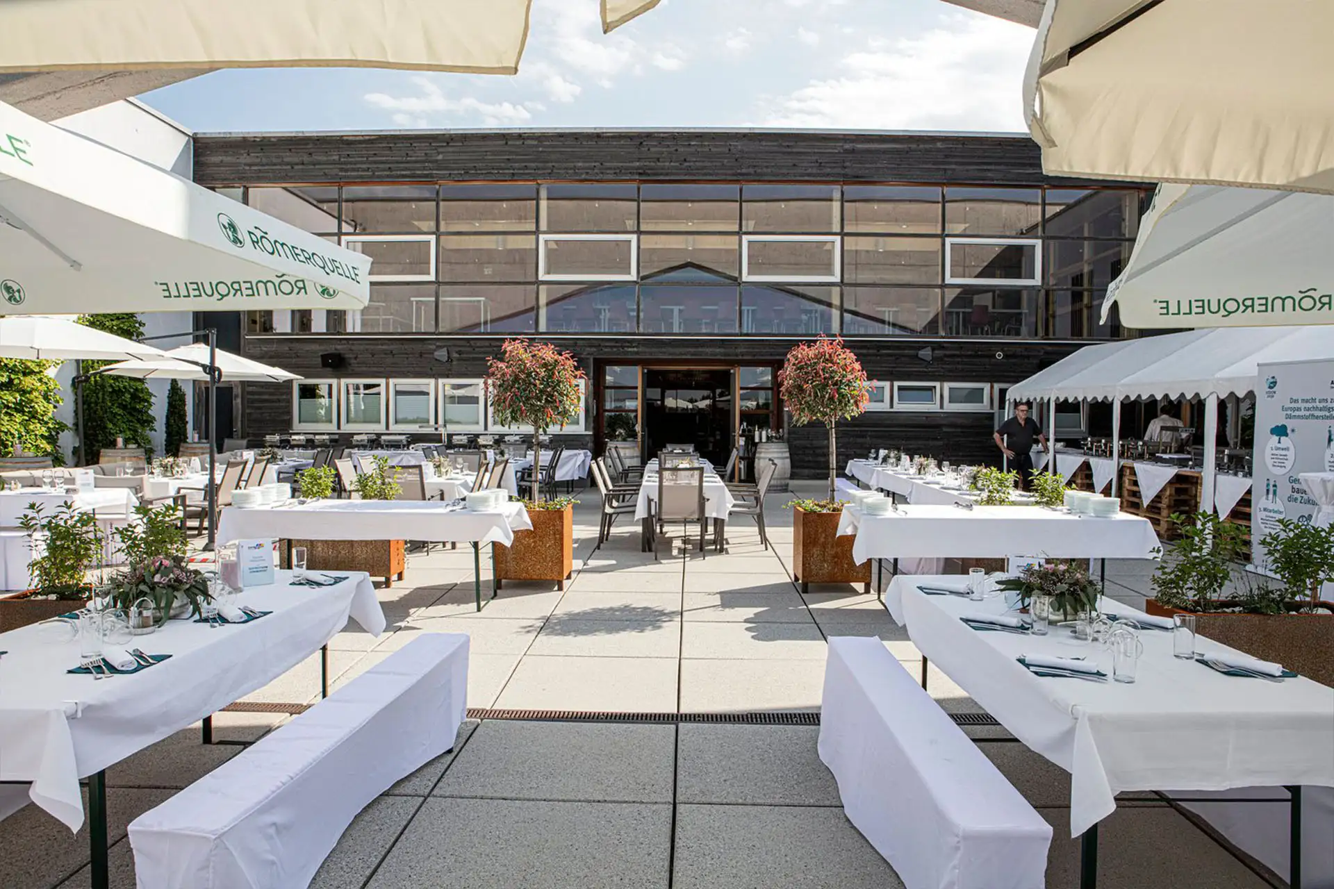 Eine Restaurantterrasse mit Tischen mit weißen Tischtüchern, Bänken, Sonnenschirmen und Topfpflanzen, die zum Essen vor einem modernen Gebäude bei teilweise bewölktem Himmel einlädt.
