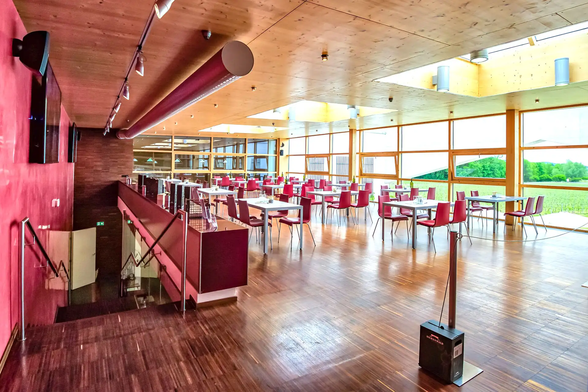 Moderne Cafeteria mit großen Fenstern, Holzböden und -decken, roten Wänden und roten Stühlen an weißen Tischen. Der Raum ist hell und bietet einen Blick auf die grüne Landschaft im Freien. Auf der linken Seite ist eine Treppe zu sehen.