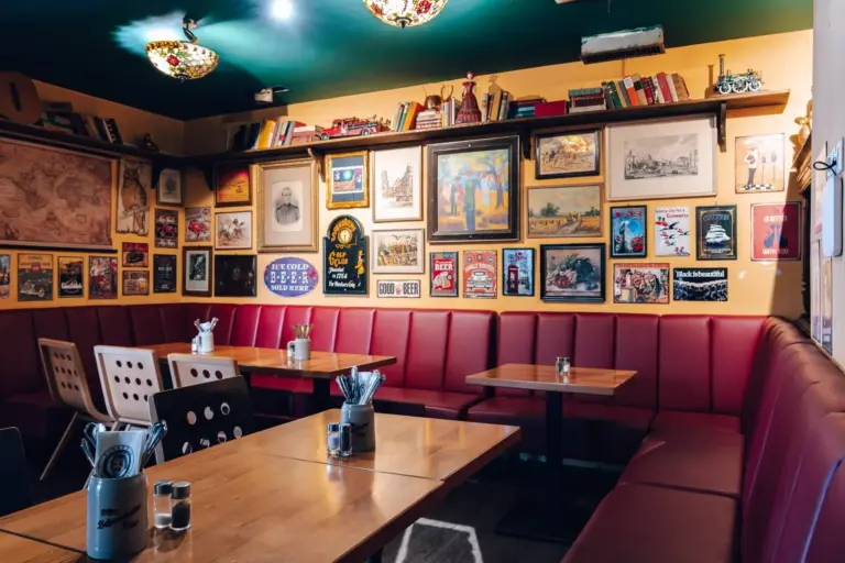 Ein gemütliches Café mit roten gepolsterten Sitzbänken, Holztischen und eklektischer Wanddekoration mit gerahmten Bildern, Landkarten und Regalen mit Büchern und alten Gegenständen.