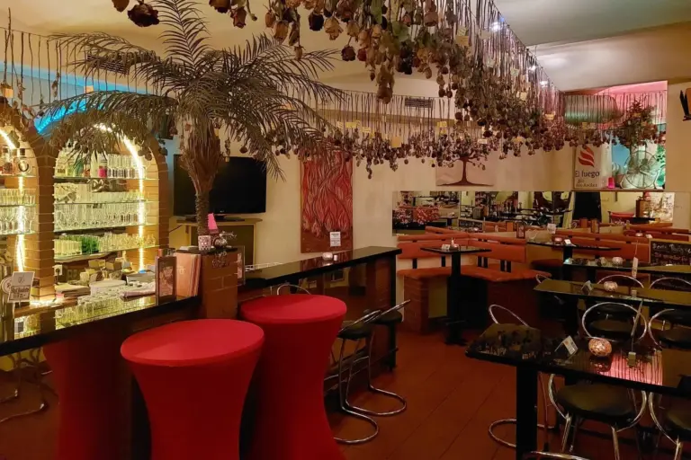 Ein gemütliches Restaurant mit Tischen, roten Hochstühlen, einer Palmen-Dekoration, hängenden Trockenblumen, einer Bar mit Gläsern und einer warmen Umgebungsbeleuchtung, die eine einladende Atmosphäre schafft.