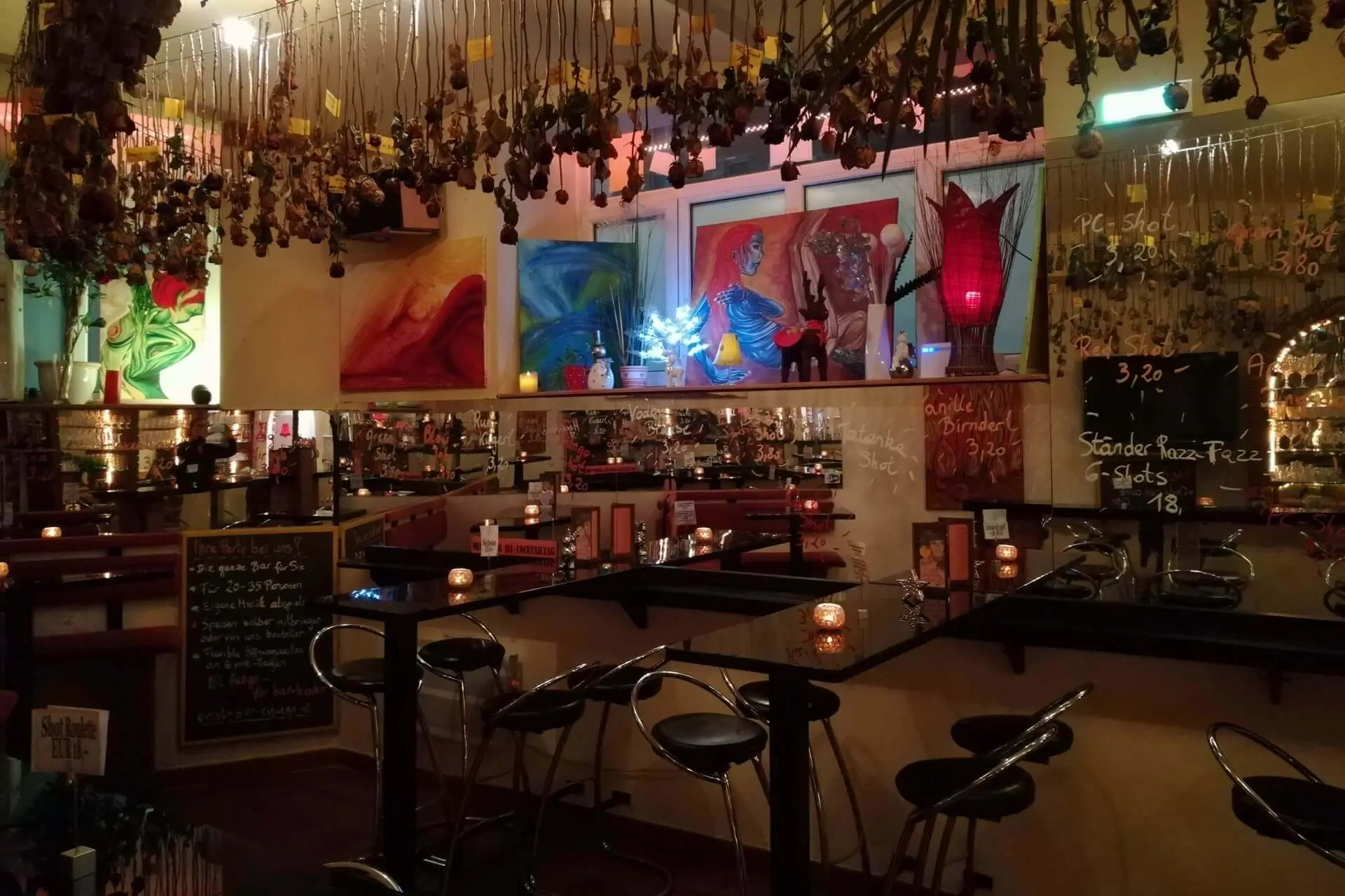 Eine gemütliche Bar mit hängenden Trockenblumen, bunten Gemälden an den Wänden, Tischen mit Kerzenlicht, Barhockern und einer Spiegelwand, die das warme, intime Ambiente widerspiegelt. Getränkespecials sind auf Tafeln geschrieben.