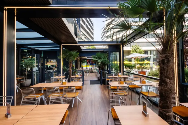 Eine moderne Restaurantterrasse mit Holztischen, Metallstühlen und getopften Palmen, umgeben von Glaswänden und hohen Gebäuden im Hintergrund an einem hellen Tag.