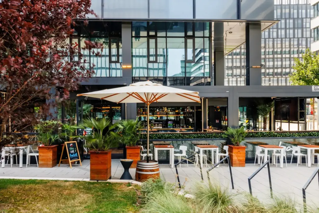 Außenterrasse eines modernen Restaurants mit Tischen und Stühlen unter einem großen weißen Sonnenschirm, umgeben von Topfpflanzen und einer Glasfassade im Hintergrund. Am Eingang befindet sich ein Schild mit der Speisekarte auf einer Kreidetafel.