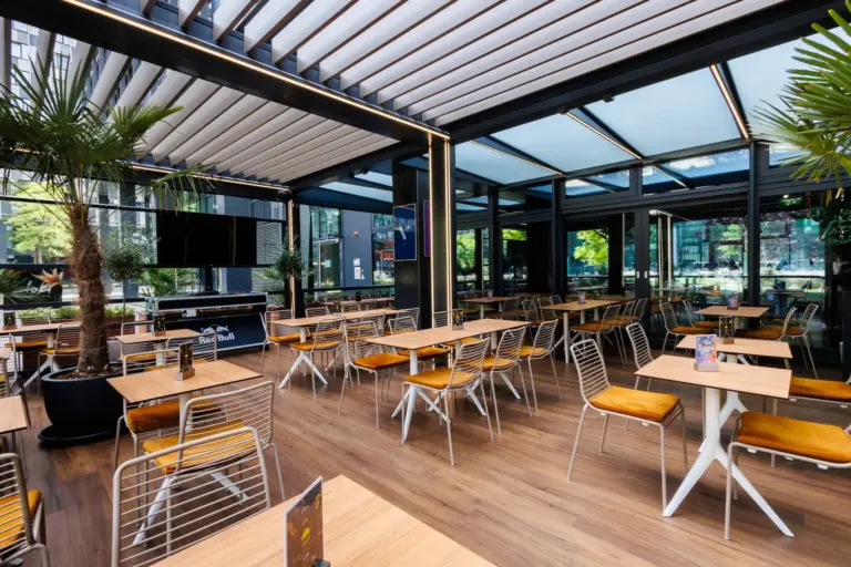 Ein modernes, geräumiges Restaurant mit Holzböden, Metallstühlen mit gelben Kissen, Holztischen, großen Topfpalmen und bodentiefen Fenstern, die viel natürliches Licht hereinlassen.