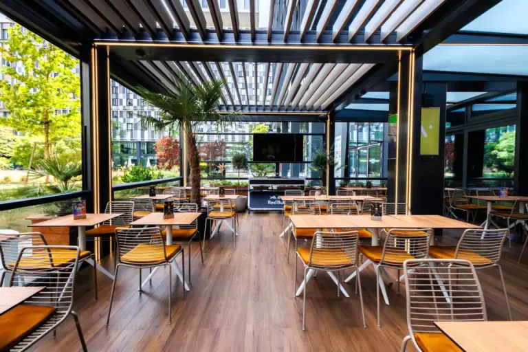 Ein modernes Freiluftrestaurant mit Holzböden, Metallstühlen und Holztischen. Große Fenster bieten einen Blick auf die grünen Bäume und Gebäude draußen. An der Rückwand ist ein Fernseher angebracht. Der Raum wirkt sauber und hell.