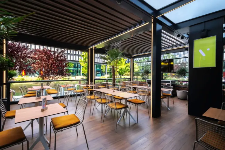 Modernes Open-Air-Café mit Holzböden, gelben gepolsterten Stühlen und quadratischen Tischen. Große Fenster und Grünflächen im Außenbereich schaffen eine helle, einladende Atmosphäre mit natürlichem Licht.