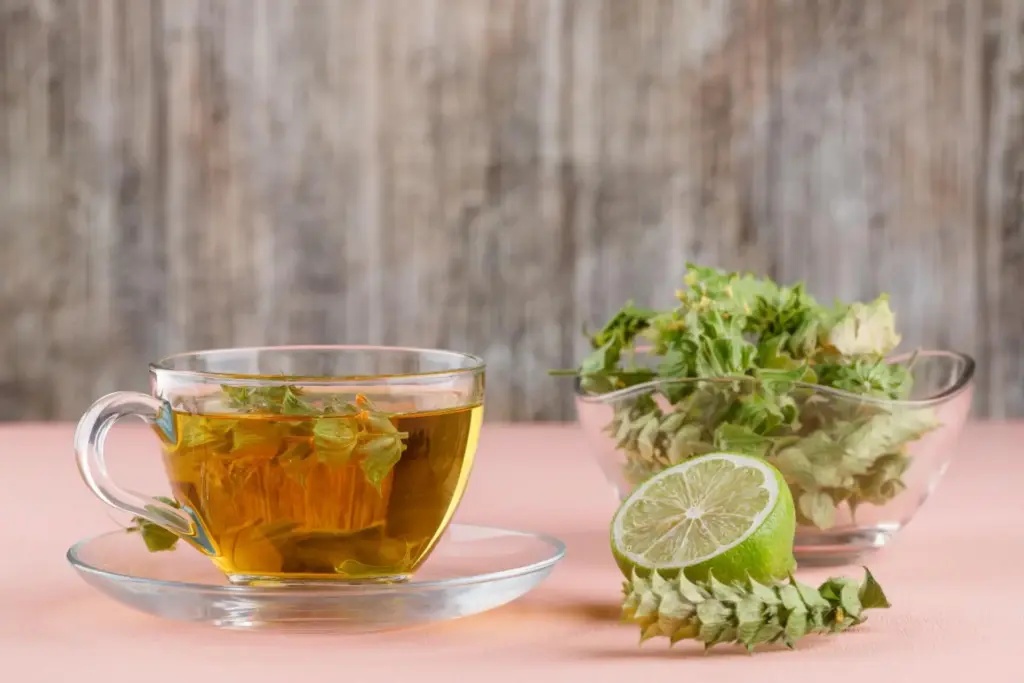 Eine Glastasse mit Kräutertee und grünen Blättern steht auf einer Untertasse, daneben eine halbierte Limette und eine Glasschale mit Kräutern, perfekt für alle, die ihre Erkältung schnell loswerden wollen, alles auf einer rosafarbenen Oberfläche mit Holzuntergrund.