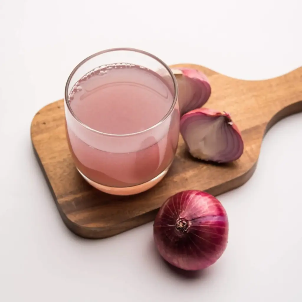 Auf einem Holzbrett steht ein Glas mit rosafarbenem Zwiebelsaft, daneben eine ganze rote Zwiebel und zwei Zwiebelhälften - ein uraltes Hausmittel, das viele Menschen verwenden, um Erkältung schnell loszuwerden, alles vor einem sauberen weißen Hintergrund.