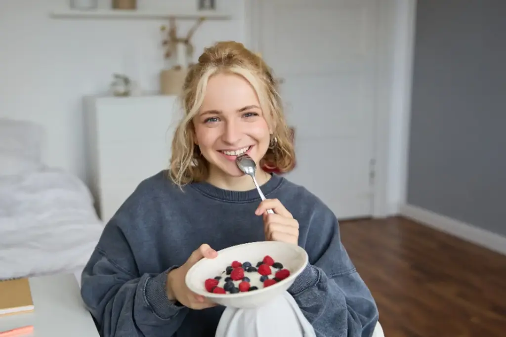 Eine lächelnde Frau mit blondem Haar und dunklem Sweatshirt sitzt in einem Raum und genießt eine Schale Joghurt mit Beeren - ein idealer Snack, um die Erkältung schnell loszuwerden. Der helle, aufgeräumte Raum hat einen Holzboden und weiße Wände.