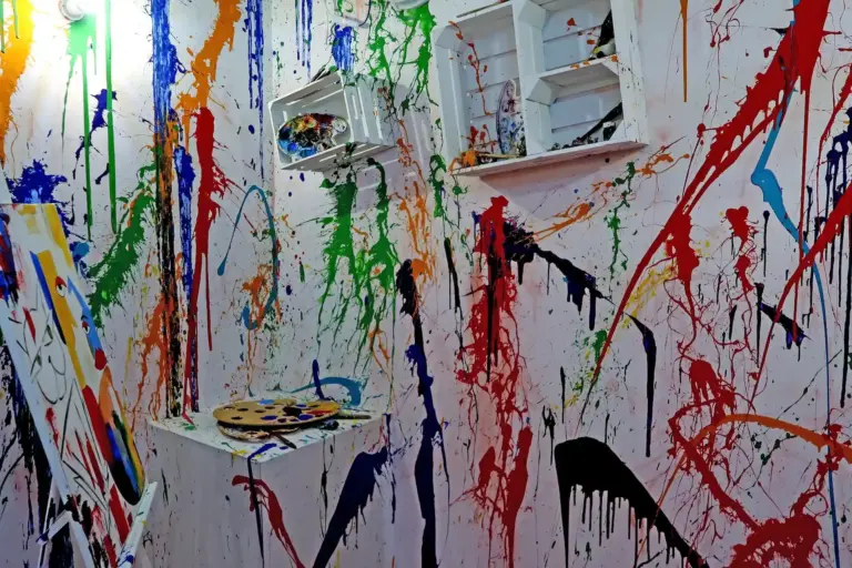 Ein weißer Raum mit Regalen, einer Palette und einer Leinwand, die mit bunten Farbspritzern und -tropfen bedeckt sind und eine chaotische, abstrakte Kunstszene ergeben.