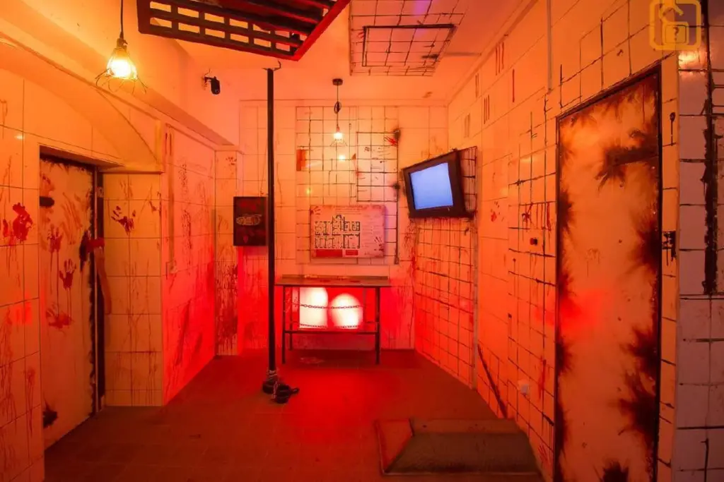 Ein schwach beleuchteter Raum mit fleckigen weißen Fliesen, roter Beleuchtung, Türen mit Rostspuren, einem schief hängenden Fernseher an der Wand und etwas, das wie Blutspritzer aussieht – all das schafft eine unheimliche, verlassene und beunruhigende Atmosphäre.