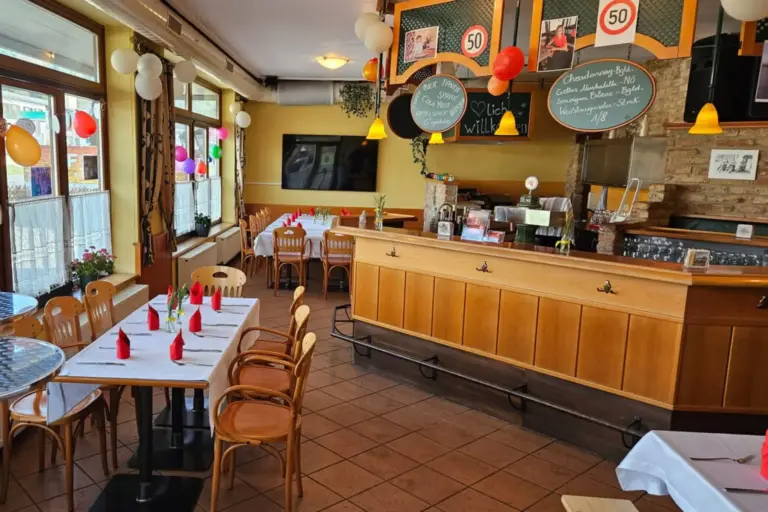 Ein gemütliches Restaurant mit Holzstühlen und Tischen mit weißen Tischtüchern und roten Servietten. Luftballons schmücken die Fenster, und im Hintergrund steht ein Tresen mit einer Kreidetafel als Speisekarte. Der Raum ist warm beleuchtet und einladend.