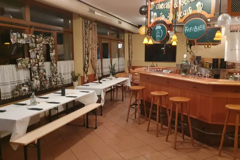 Ein gemütliches Restaurant mit Holzmöbeln, langen Tischen mit weißen Tischdecken, Bänken, Barhockern, einem dekorierten Barbereich und festlicher Dekoration an der Decke. An der Wand sind Fotos ausgestellt.