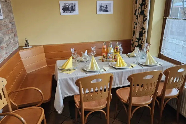Ein Tisch in einem gemütlichen Restaurant ist für sechs Personen gedeckt, mit gelben gefalteten Servietten, Gläsern, Getränken und Stühlen. Ein Fenster und eine Wand mit gerahmten Fotos schaffen eine warme, einladende Atmosphäre.