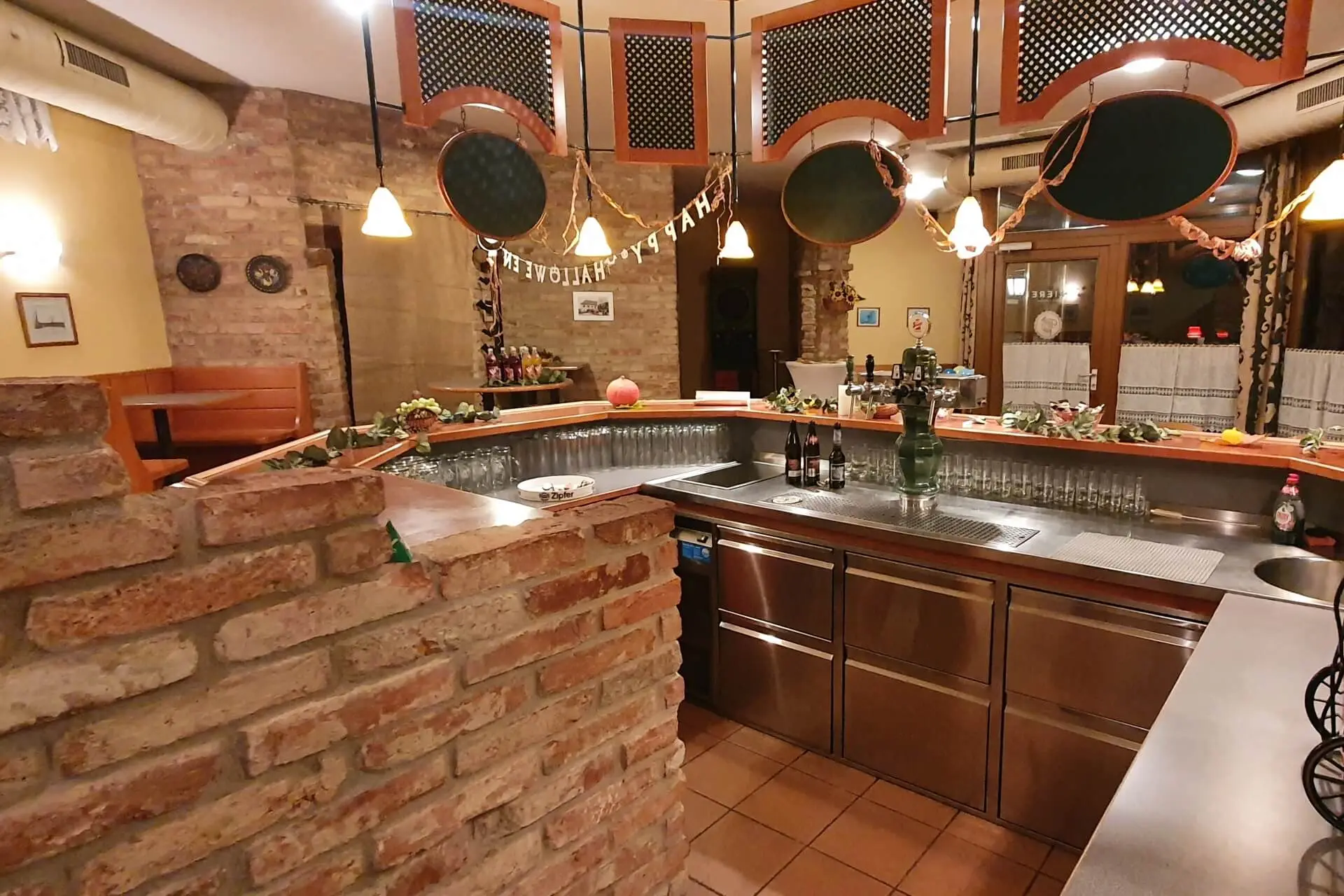 Ein gemütlicher Barbereich mit Backstein- und Edelstahloberflächen, Hängelampen, dekorativer Girlande und mehreren Flaschen und Gläsern auf dem Tresen. Im Hintergrund sind Tische und Stühle zu sehen.